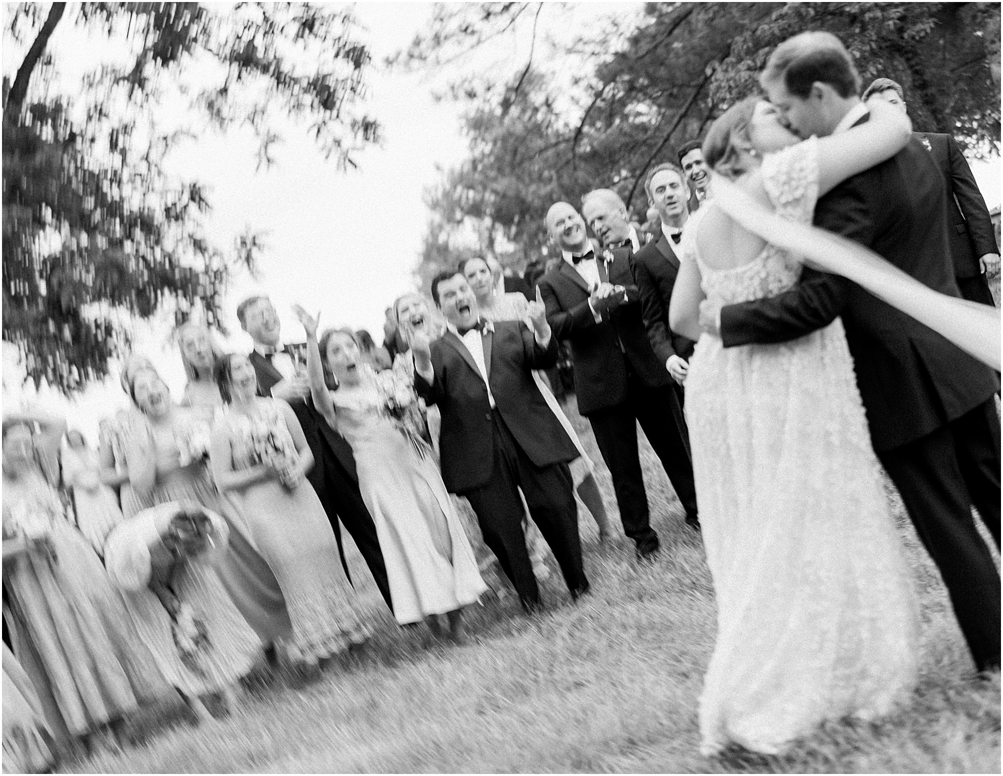Bride and groom kissing infront of wedding party as everyone cheers. Black and white blurry photo of bride and groom with wedding party.